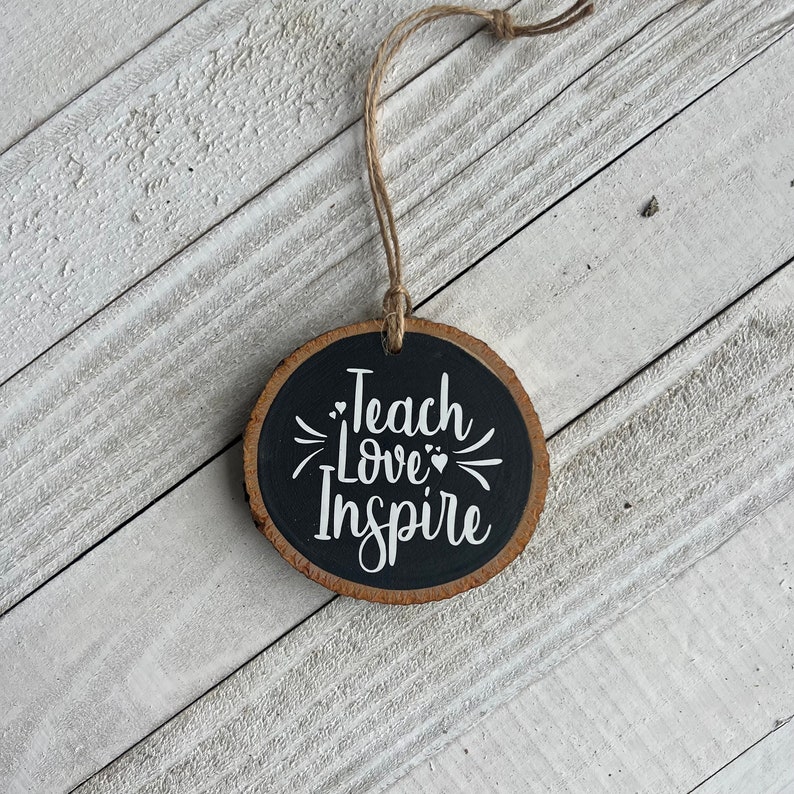 May include: A round wooden ornament with a black painted surface. The ornament has a white string for hanging and the words "Teach Love Inspire" written in white on the front.
