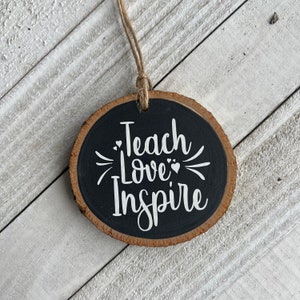 May include: A round wooden ornament with a black background and white text that reads "Teach Love Inspire". The ornament has a brown wood grain edge and a twine string for hanging.