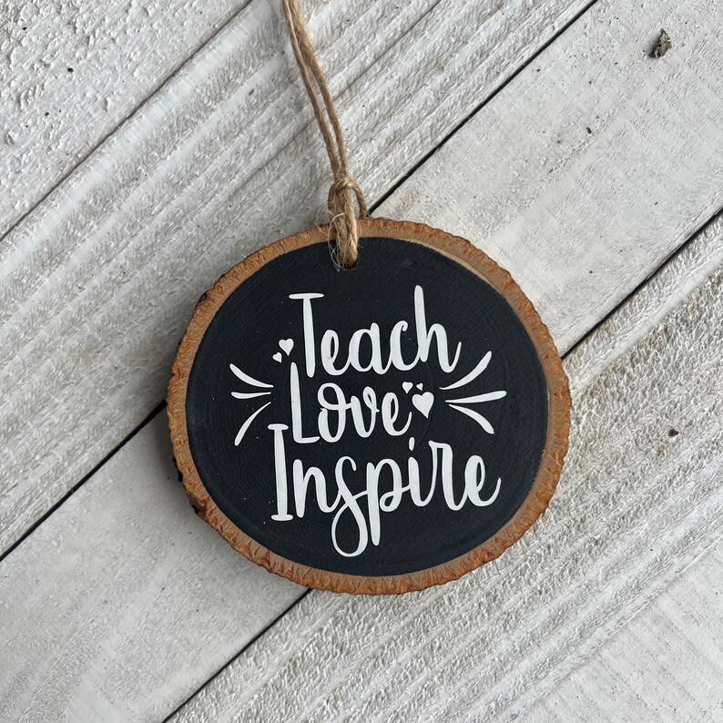 May include: A round wooden ornament with a black background and white text that reads "Teach Love Inspire". The ornament has a brown wood grain edge and a twine string for hanging.