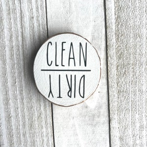 May include: A round wooden sign with the words "CLEAN" and "DIRTY" written on it in black paint. The sign is white and has a rustic look.