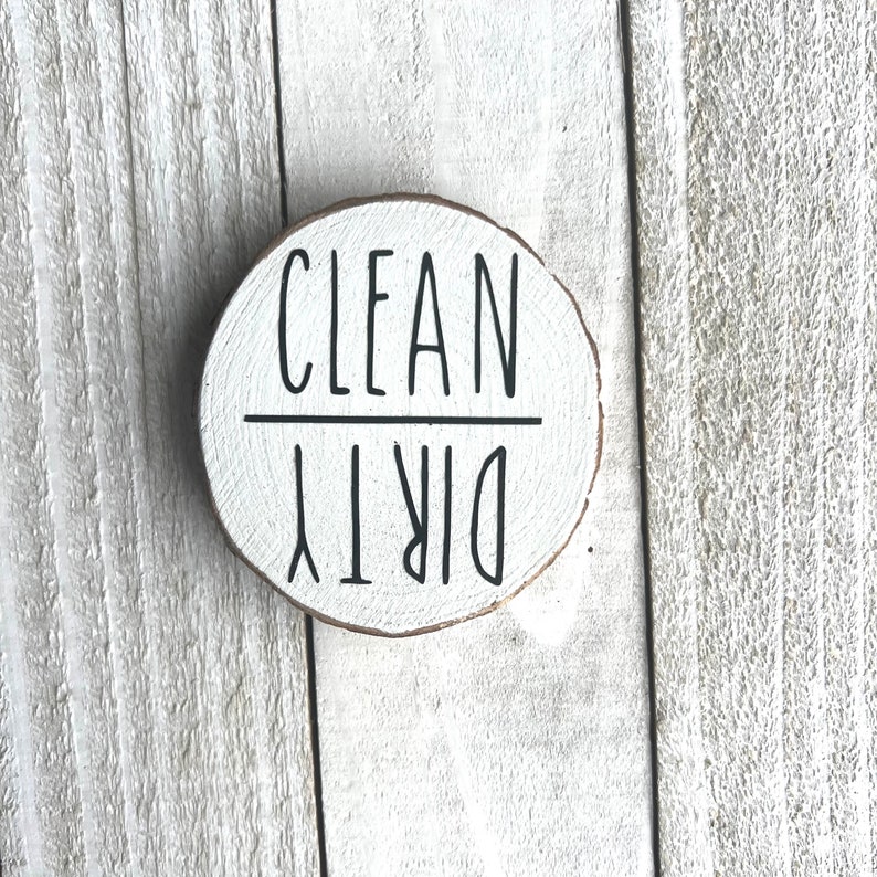 May include: A round wooden sign with the words "CLEAN" and "DIRTY" written on it in black paint. The sign is white and has a rustic look.