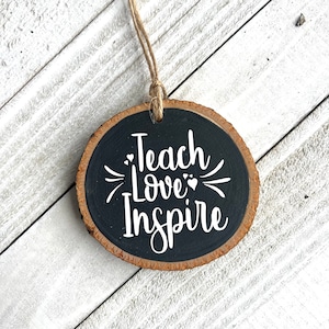 May include: A round wooden ornament with a black background and white text that reads "Teach Love Inspire".