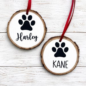 May include: Two round wooden slice ornaments with natural bark edges, each featuring a white painted center. One ornament displays a black paw print and the name Harley in script, while the other shows a black paw print and the name KANE in sans-serif.