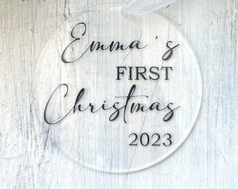 Puede incluir: Adorno redondo transparente con texto negro que dice "Emma's First Christmas 2023".