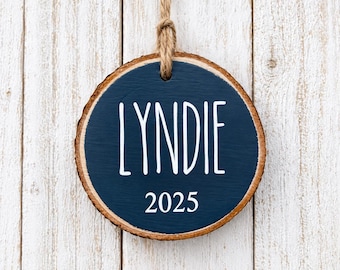 May include: A round wooden slice ornament with natural bark edges, featuring a dark navy blue painted center. The name LYNDIE is printed in white sans-serif capital letters above the year 2025. A brown jute twine loop is attached for hanging.