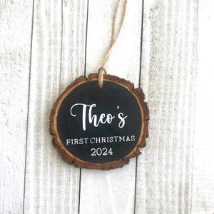 May include: A round wooden ornament with a black painted surface. The ornament has the text "Theo's First Christmas 2024" written in white paint.