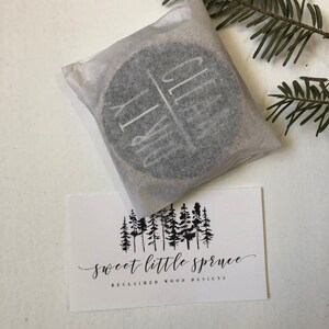 May include: A round wooden coaster with the words "Dirty" and "Clean" written on it. The coaster is wrapped in white tissue paper and is sitting on a white surface. The image also includes a business card with the text "sweet little spruce reclaimed wood designs" and a graphic of a row of pine trees.