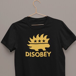 May include: Black t-shirt with a gold graphic of a hedgehog-like creature with three stars above it and the word "DISOBEY" below. The shirt is hanging on a gold hanger against a gray background.