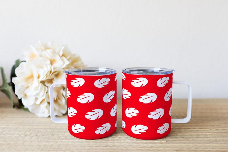 May include: Two red travel mugs with white leaf patterns. Each mug has a white handle and a clear lid. The mugs are on a wooden surface with a white flower arrangement in the background.