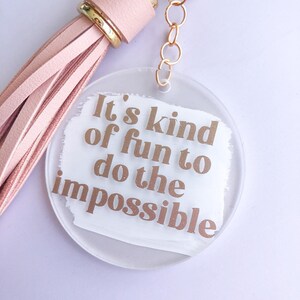 May include: A white round acrylic keychain with a pink tassel. The keychain has the text "It's kind of fun to do the impossible" in gold glitter lettering.