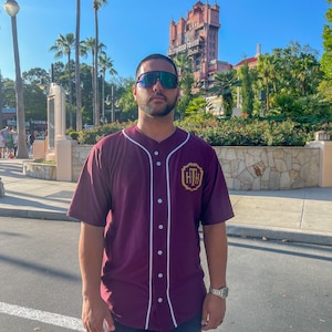 May include: A short-sleeved, button-down baseball jersey in a deep maroon color. The shirt features white piping along the front placket and a gold-colored emblem on the left chest with the letters HTH inside a decorative border design.