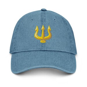May include: A light blue denim baseball cap with a gold trident embroidered on the front. The cap has a curved brim and an adjustable strap at the back. The trident design is a symbol often associated with mythology and power.