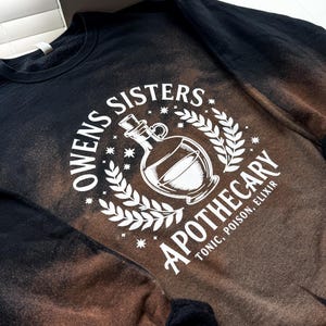 May include: Black and brown bleached sweatshirt with the text "Owens Sisters Apothecary" in a white circular design. The design includes a potion bottle, stars, and a laurel wreath. The sweatshirt reads "Tonic, Poison, Elixir."