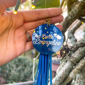 May include: A circular dark blue acrylic keychain featuring the white text Ma Belle Evangeline and a small white firefly graphic. The keychain is attached to a gold-toned metal ring and includes a long, bright blue faux leather tassel fringe.