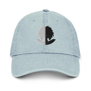 May include: A light blue denim baseball cap with a black and white embroidered design of a stylised head with a half-circle shape.