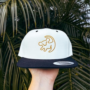 Puede incluir: Gorra de béisbol blanca y negra con un contorno bordado en oro de Simba de El Rey León.