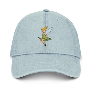 May include: Light blue denim baseball cap featuring an embroidered image of a fairy in a green dress and wings. The fairy is sitting on a green leaf. The cap has a curved brim and a fabric strap closure.
