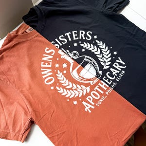May include: Two t-shirts, one black and one orange, with white graphic designs. The black shirt features the text "SISTERS APOTHECARY" around a bottle illustration. The orange shirt has the text "OWENS SISTERS APOTHECARY" with a similar design.