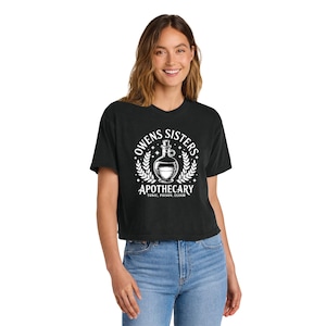 May include: Black cropped t-shirt with a white graphic that reads "Owens Sisters Apothecary" with a bottle illustration. The shirt is paired with blue jeans. The design includes a bottle, laurel leaves, and the words "Tonic, Poison, Elixir."