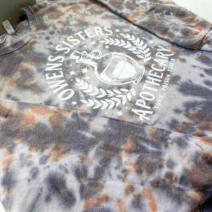 May include: A tie-dye long-sleeve shirt in shades of gray, orange, and white. The shirt features the text "Owens Sisters Apothecary" around a graphic of a potion bottle. The text "Tonic, Poison, Elixir" is also printed on the shirt.