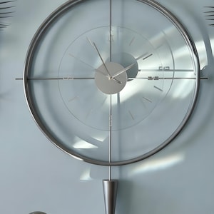 May include: A modern wall clock with a clear glass face and a silver metal frame. The clock features a circular design with minimalist hour markers and hands. A silver pendulum hangs below the clock face. The clock is mounted on a light blue wall.