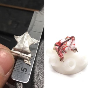 May include: Two origami frogs, one white and one with a red and white floral pattern. The white frog is being held in a hand, while the patterned frog is sitting on a white base.