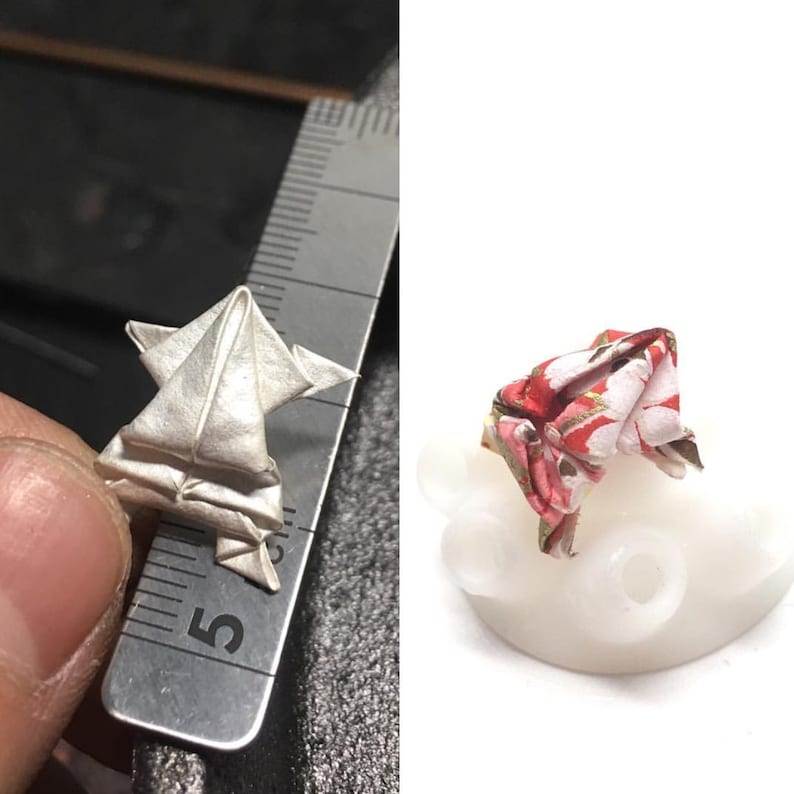 May include: Two origami frogs, one white and one with a red and white floral pattern. The white frog is being held in a hand, while the patterned frog is sitting on a white base.