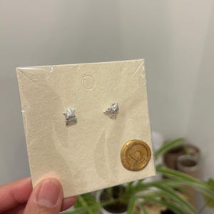 May include: A pair of silver geometric stud earrings displayed on a beige card with a gold wax seal.
