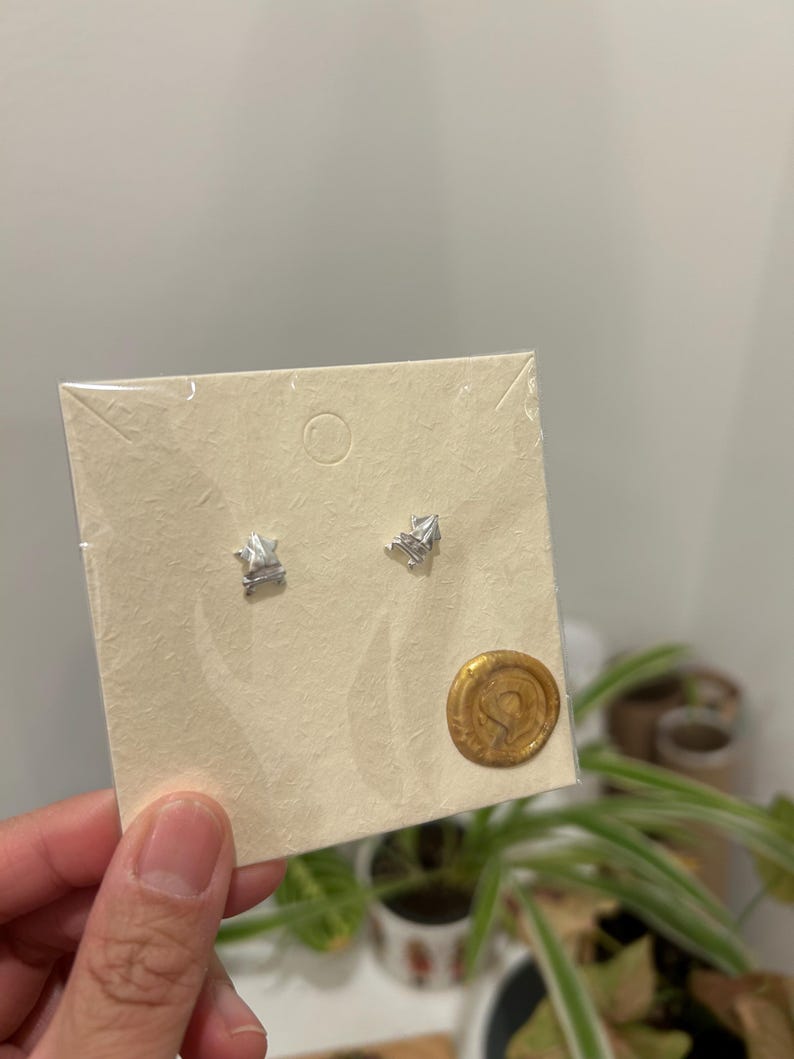 May include: A pair of silver geometric stud earrings displayed on a beige card with a gold wax seal.