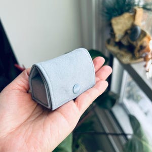 May include: A small, light blue jewelry box with a rounded top and a button closure. The box is made of a soft, felt-like material. The jewelry box is held in an open hand.