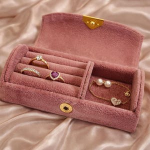 May include: A pink velvet jewellery box with gold hardware, open to reveal rings and a bracelet. The rings include a diamond-like flower, a curved band of small stones, and a heart-shaped ruby. The bracelet has pearls and a heart charm.