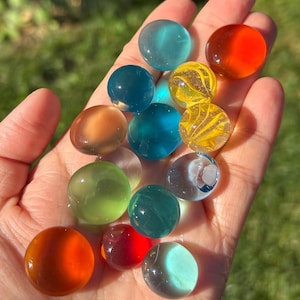 Peut inclure: Un ensemble de billes de verre colorées dans diverses nuances de bleu, vert, orange et jaune. Les billes sont tenues dans une main, la lumière du soleil soulignant leur qualité translucide. Les billes sont un jouet classique.