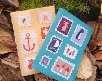 Può includere: Due quaderni in tessuto di feltro con disegni ricamati colorati. I quaderni sono decorati con varie immagini, tra cui un'ape, una stella, un'ancora, un coniglio, un fungo, un pinguino e un rametto di lavanda. I quaderni sono di colore giallo chiaro e blu turchese.