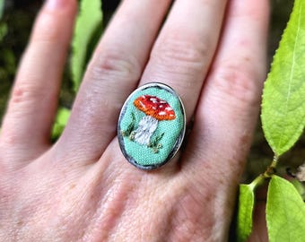 Può includere: Un anello d'argento con una base in tessuto verde e un design di fungo rosso e bianco ricamato.