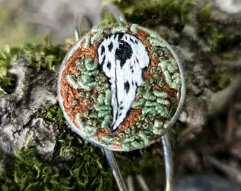 Può includere: Un braccialetto d'argento con un ciondolo rotondo ricamato che presenta un design di uccello bianco e nero su uno sfondo verde e marrone.