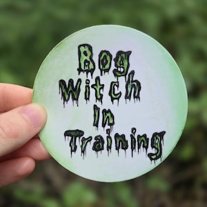 May include: A white round sticker with a green and black dripping font that reads "Bog Witch in Training".