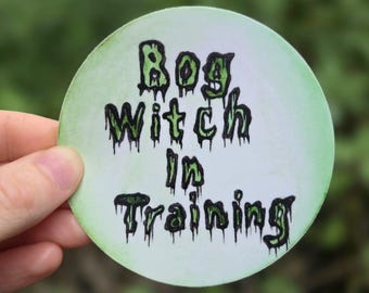 Può includere: Un adesivo rotondo bianco con un font verde e nero che cola e che dice "Bog Witch in Training".