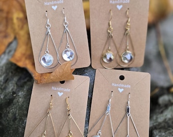 Può includere: Quattro paia di orecchini fatti a mano esposti su cartoncini marroni. Gli orecchini presentano pendenti di cristallo trasparente e rotondo e sono disponibili in design in metallo argentato e dorato, tra cui forme a goccia e geometriche. Le carte hanno la parola "Handmade" stampata.