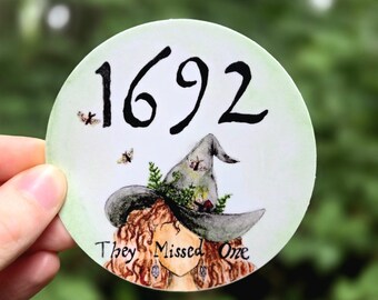 Può includere: Un adesivo rotondo con una strega che indossa un cappello verde e grigio con il numero 1692 e il testo "They Missed One".
