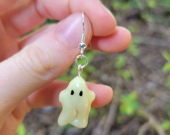 Può includere: Un paio di orecchini pendenti d'argento con un piccolo ciondolo a forma di fantasma giallo chiaro. Il fantasma ha occhi neri e una forma semplice e arrotondata.