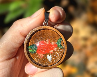 Può includere: Ciondolo rotondo in legno con un design a pesce rosso e dettagli verdi su sfondo marrone. Il ciondolo è tenuto in mano, con uno sfondo sfocato di fogliame autunnale.