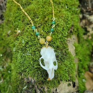 May include: A gold-colored chain necklace featuring a small animal skull pendant. The necklace includes green and yellow beads. The skull is white and tan, and the chain is draped over green moss.