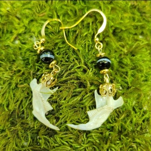 May include: Gold-colored dangle earrings featuring a claw-shaped bone pendant. Each earring has a black and white striped bead and a small gold-colored crown-like charm. The earrings are displayed on a bed of green moss.