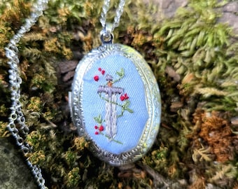 Può includere: Collana con ciondolo ovale color argento. Il ciondolo presenta un ricamo di una croce con bacche rosse e foglie verdi su sfondo azzurro. La collana è esposta su muschio verde.