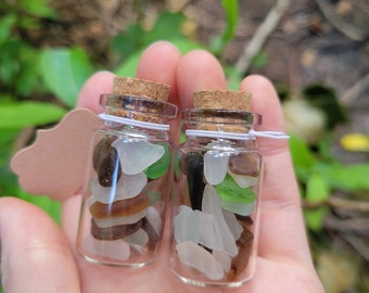 Può includere: Due piccole bottiglie di vetro con tappi di sughero riempite di vetro di mare colorato. Le bottiglie sono tenute nella mano di una persona.