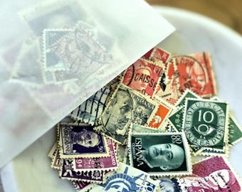 Può includere: Una collezione di francobolli d'epoca fuoriesce da una busta di carta traslucida in una ciotola bianca. I francobolli presentano vari colori, ritratti e testo, tra cui "USA 16c" e "ESPANA CORREOS".