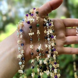 May include: Several strands of necklaces with bone-shaped beads and colorful gemstone beads in shades of purple, green, blue, and black. The necklaces are held up against a blurred green background, showcasing the jewelry's intricate design and color variations.