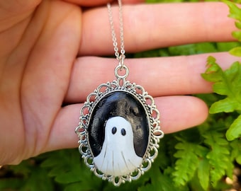 Può includere: Una collana d'argento con un ciondolo a forma di fantasma bianco e nero. Il fantasma è dipinto su uno sfondo nero e ha due occhi bianchi. Il ciondolo è circondato da una cornice in filigrana d'argento.