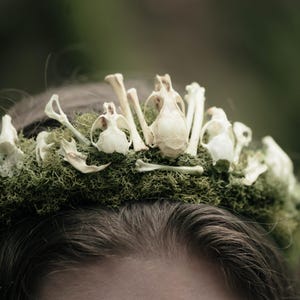 Puede incluir: Una corona única con una base de musgo verde adornada con pequeños cráneos y huesos de animales blancos. La corona se asienta sobre el cabello oscuro de una persona, con un fondo verde borroso.