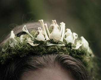 Può includere: Un'originale corona con una base di muschio verde ornata da piccoli teschi e ossa di animali bianchi. La corona è appoggiata sui capelli castano scuro di una persona, con uno sfondo verde sfocato.
